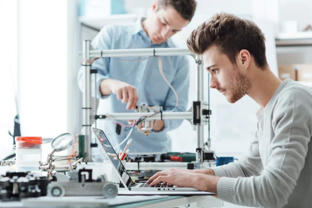 Zwei Ingenieure entwickeln Elektronik: Einer am Laptop, der andere am 3D-Drucker.
